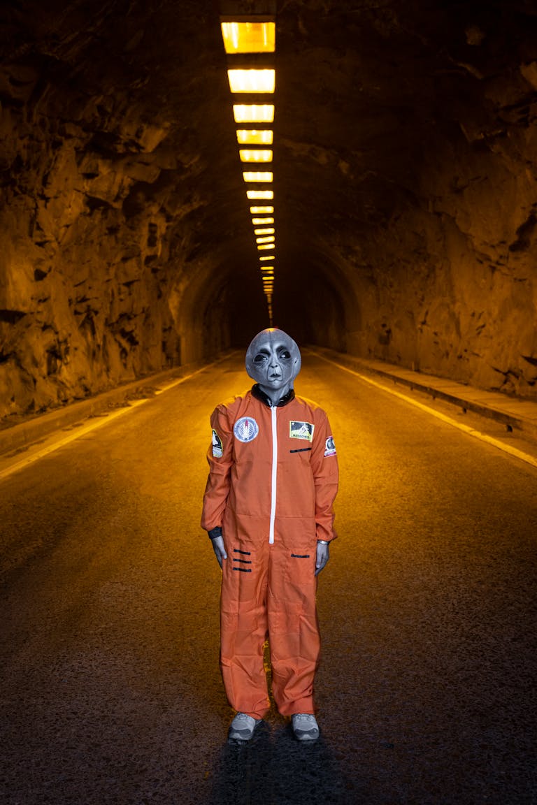A grey alien posing in an orange astronaut suit inside an illuminated tunnel.
