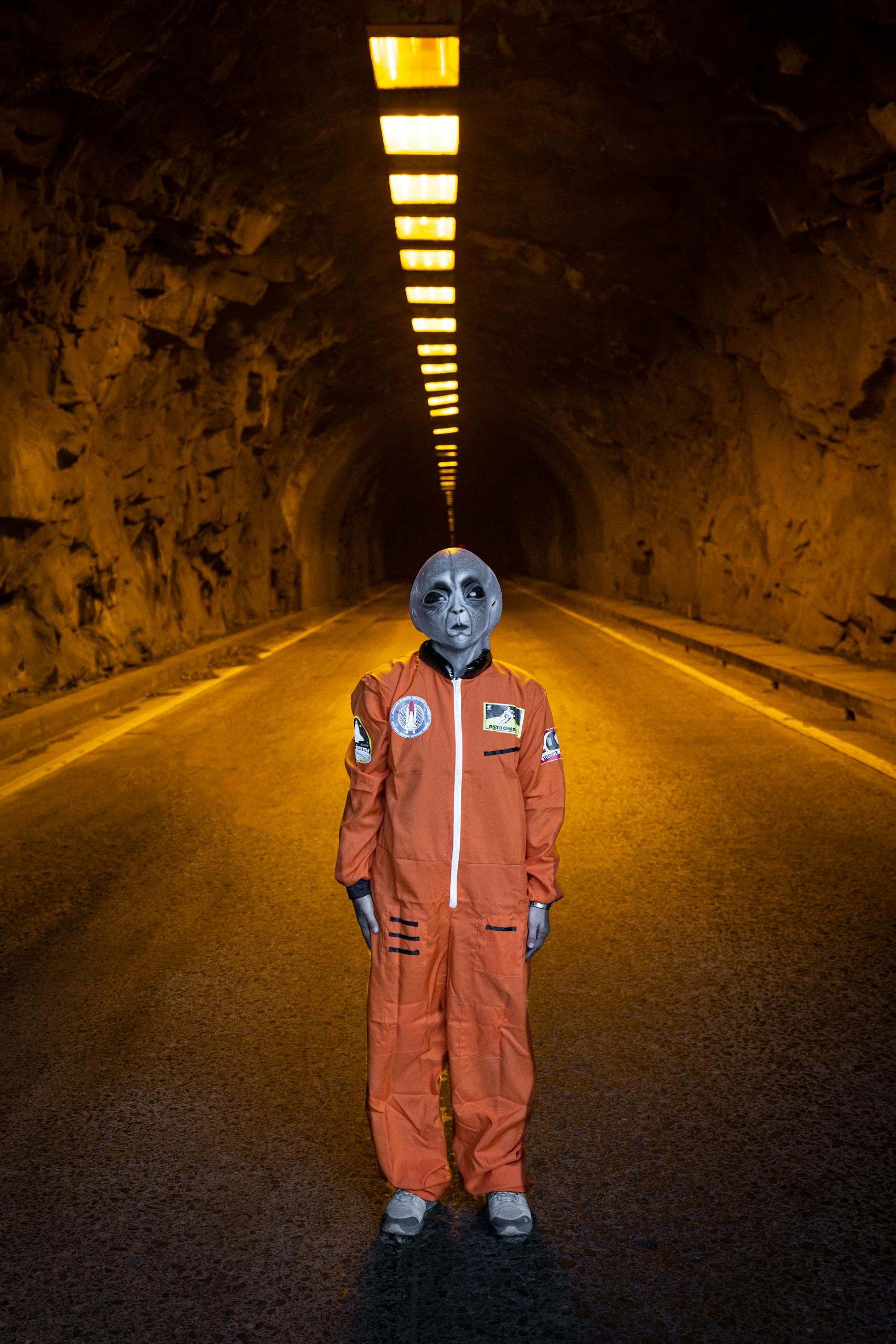 A grey alien posing in an orange astronaut suit inside an illuminated tunnel.